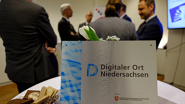 Auf einem Tisch im Vordergrund steht ein Schild mit der Aufschrift „Digitaler Ort Niedersachsen“, im Hintergrund unterhalten sich sechs Personen. 